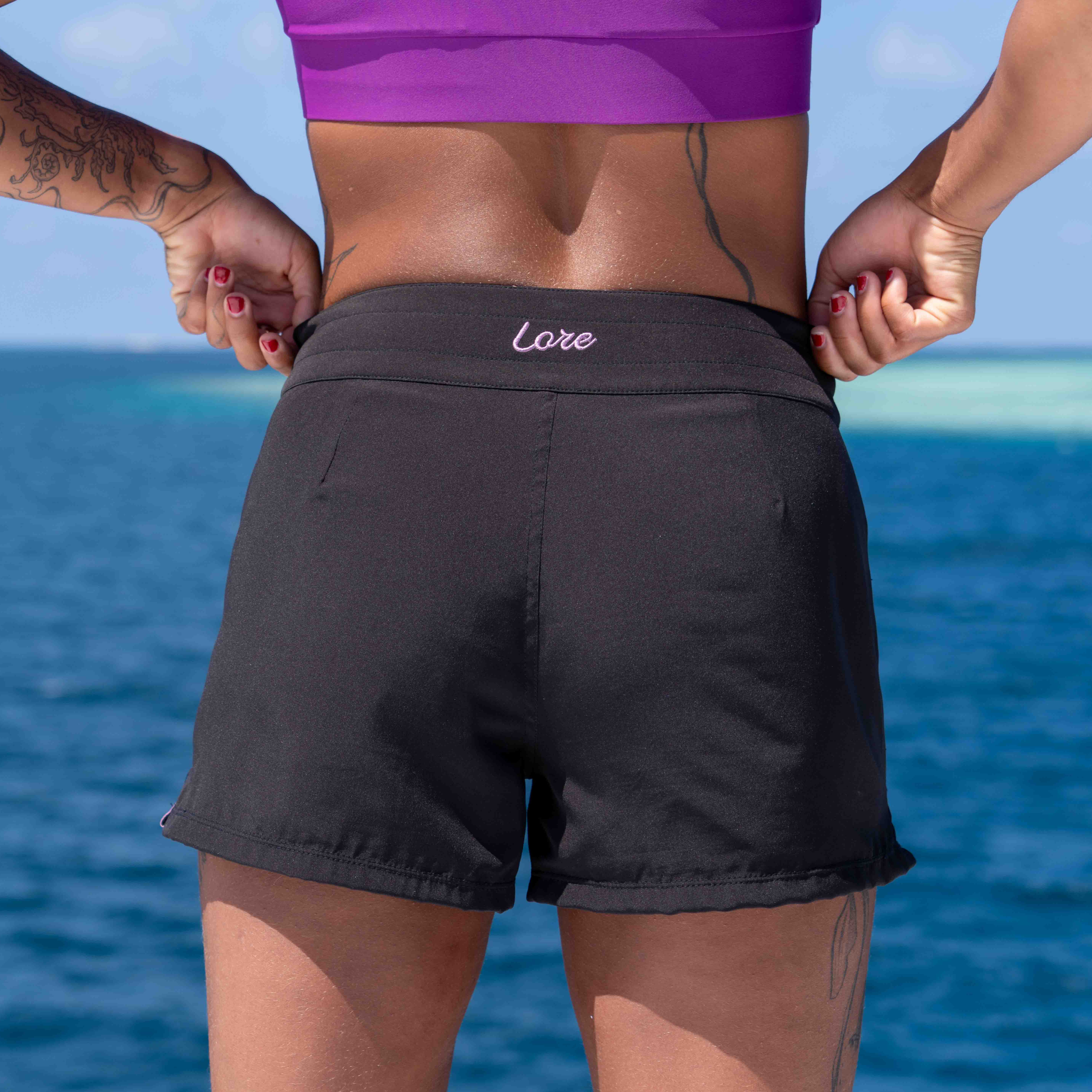 Person wearing black shorts with 'Lore' embroidery against a blue ocean background