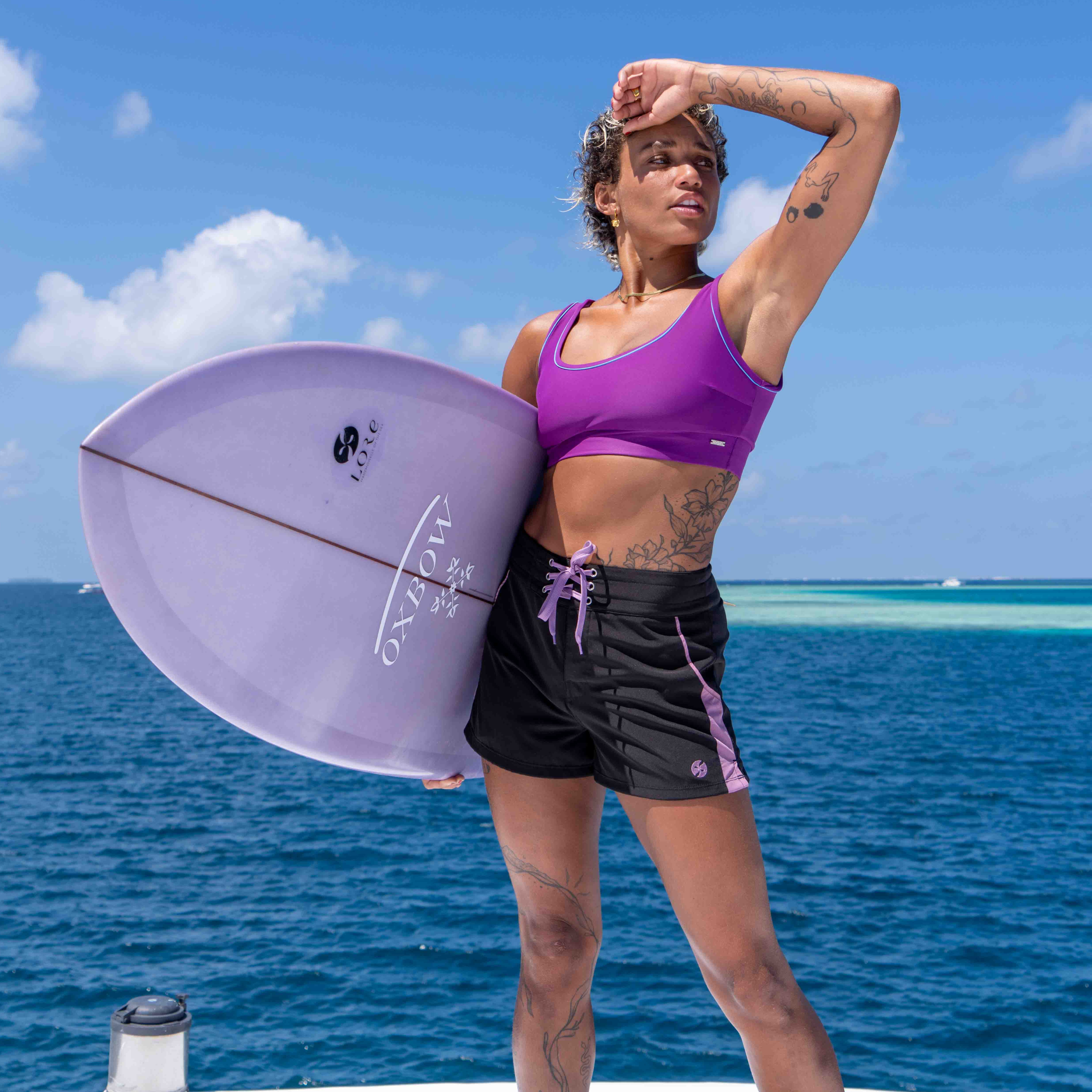 Woman in bikini top and black shorts holding a purple surfboard with ocean and sky in the background